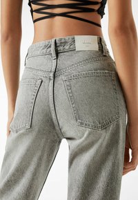 Parte baja de la espalda y caderas de una persona que lleva puestos unos jeans de mezclilla grises de tiro alto con bolsillos traseros y un top corto negro con diseño entrecruzado.