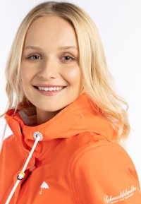 Ljusorange vattentät jacka med hög krage, vita detaljer och en logotyp med en vit paraply. Slät textur och dragsko.