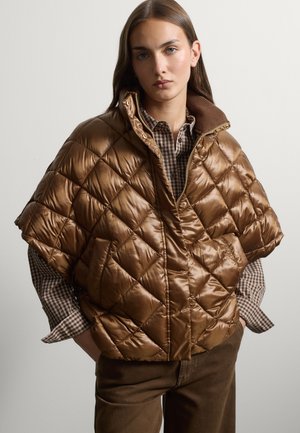 Woman wearing a shiny brown quilted short-sleeve jacket over a checkered shirt and brown pants, standing with hands in pockets.