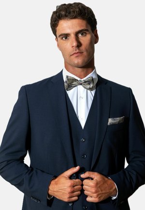 Man dressed in a dark blue three-piece suit with a white shirt and patterned bow tie, adjusting jacket buttons against a plain background.