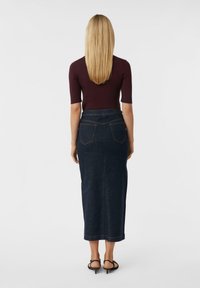 Frau steht und trägt ein dunkelviolettes kurzärmeliges Oberteil, einen dunklen Jeans-Midirock mit Rückentaschen und schwarze High Heels, vor einem weißen Hintergrund.