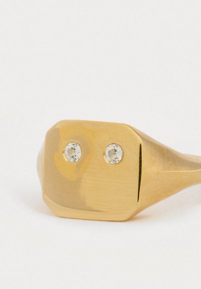 Zlatý pečetní prsten s dvěma malými průhlednými drahokamy umístěnými vedle sebe na plochém, leštěném obdélníkovém poli na bílém pozadí.