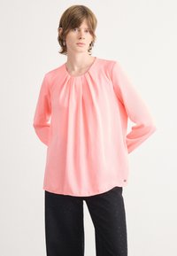 Rosa Bluse mit langen Ärmeln, gerüschtem Ausschnitt und weicher, glatter Textur; kombiniert mit dunklen Hosen, die am Saum dezente Glitzerakzente haben.