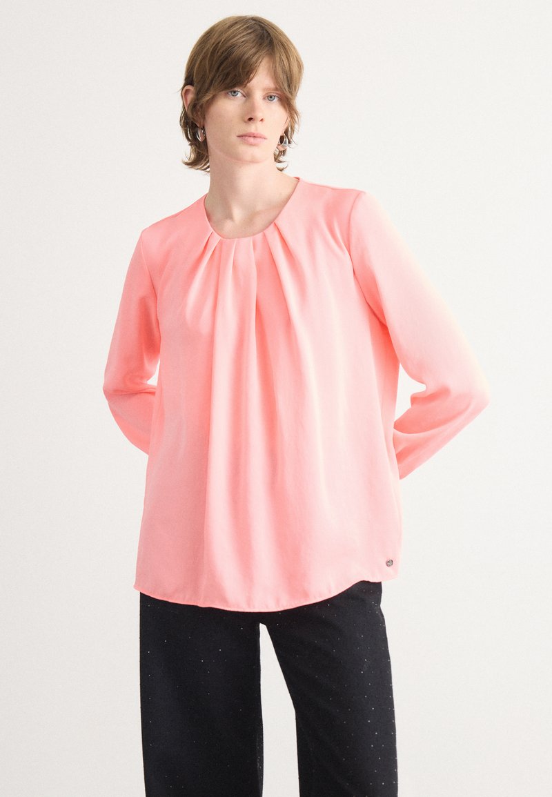 Rosa Bluse mit langen Ärmeln, gerüschtem Ausschnitt und weicher, glatter Textur; kombiniert mit dunklen Hosen, die am Saum dezente Glitzerakzente haben.