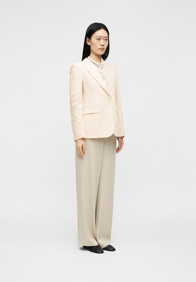 Kvinde stående iført en cremefarvet blazer, beige bukser med vide ben, cremefarvet top og sorte spidse sko mod en ensfarvet lys baggrund.