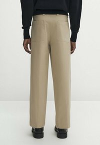 Person wearing beige straight-leg pants with back pockets, a black long-sleeve top, and black shoes, standing against a plain background.