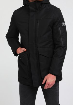 Mand iført sort isoleret jakke med hætte, flere lynlåslommer og et rundt logo på venstre ærme.