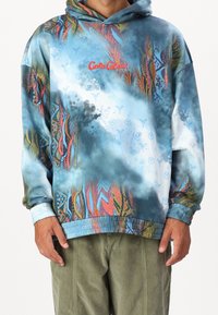 Uomo che indossa un hoodie blu con motivo multicolore e la scritta "Cools Colucci" in rosso, insieme a pantaloni verde oliva, in piedi su uno sfondo bianco.