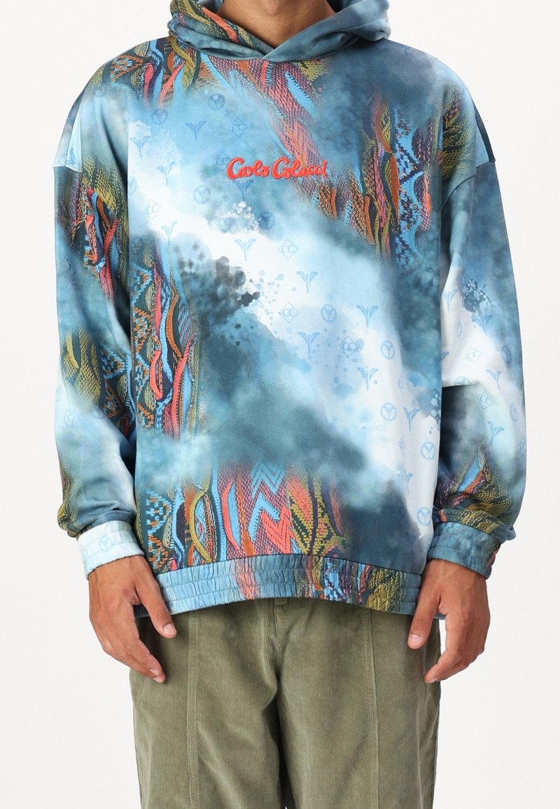 Uomo che indossa un hoodie blu con motivo multicolore e la scritta "Cools Colucci" in rosso, insieme a pantaloni verde oliva, in piedi su uno sfondo bianco.