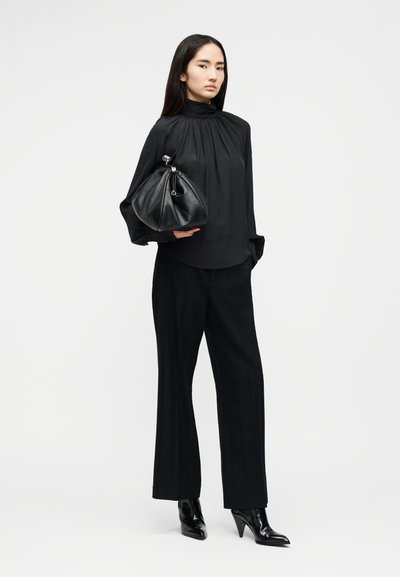 Femme aux longs cheveux noirs portant un chemisier noir ample, un pantalon noir à jambes larges, des bottes à talons noires, tenant un grand sac en cuir noir.