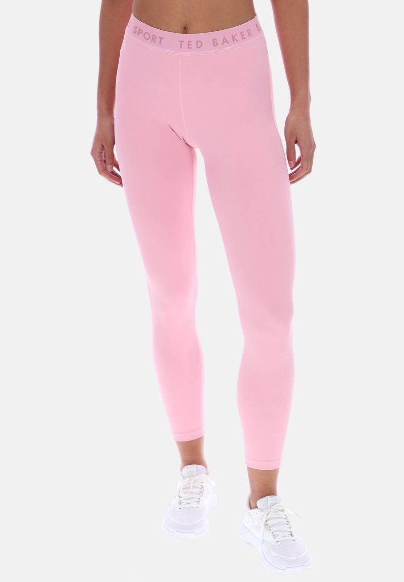 Femme portant un legging de sport Ted Baker rose clair et des baskets blanches, debout devant un fond blanc uni.