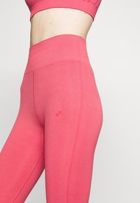 Dopasowane różowe legginsy wykonane z gładkiego, elastycznego materiału z wysokim stanem i subtelnym detalem teksturowym po bokach. Zawierają małe logo na udzie.