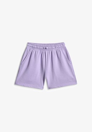 Fille debout avec un t-shirt rose oversize, un short lilas, des baskets assorties, des chaussettes blanches et un sac à épaule lilas sur un fond gris.