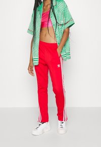 Rote Jogginghose mit weißen Seitstreifen, pinkes Cropped-Top und eine grün gemusterte kurzärmlige Jacke. Weiße Sneakers runden das Outfit ab.