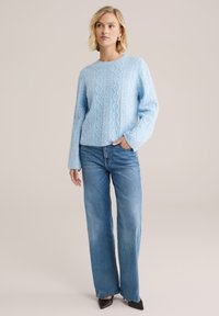 Pulover tricotat de culoare bleu deschis, cu un model cu cabluri, decolteu rotund și mâneci lungi, purtat cu blugi bleu cu talie înaltă și croială largă.