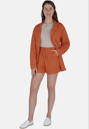 Oranges Leinenhemd und passendes Shorts-Set, das einen lockeren Schnitt, einen elastischen Bund und ein helles neutrales Oberteil darunter bietet. Weiße Slipper.