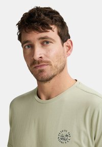 Hellgrünes Baumwoll-T-Shirt mit Rundhalsausschnitt, das ein kleines schwarzes rundes Logo auf der linken Brust und eine glatte Textur aufweist.