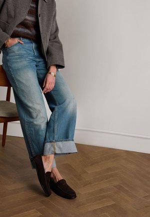 Lichtblauwe wijde jeans met een opgerolde zoom, gedragen met donkerbruine suède instappers. Een grijze jas en een gebreide trui zijn ook zichtbaar.
