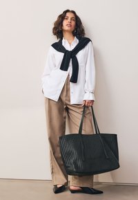 Sac cabas tissé noir avec de longues poignées, associé à une chemise blanche boutonnée, un pantalon beige taille haute et des mules noires, posé devant un fond clair.