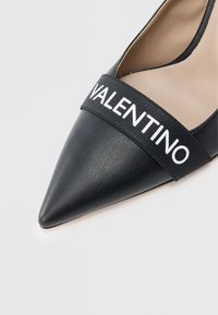 Czarny skórzany but z ostrym noskiem i szeroką czarną elastyczną opaską z białym napisem "VALENTINO" na przodzie. Gładka tekstura.