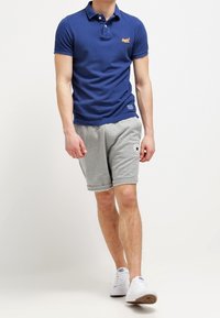 Mörkblå polo med en logotyp på bröstet, matchad med ljusgrå shorts. Shortsen har uppvikta fållar och en rund logotyp-patch. Vita sneakers.