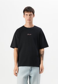 Young man with short dark hair wearing a black oversized T-shirt with "Turbo" text and light blue jeans, standing against a white background.
