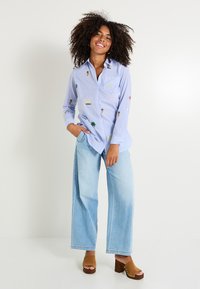 Camisa de rayas azules con bordados coloridos de iconos como piñas y arcoíris; combinada con vaqueros de pierna ancha en azul claro y zuecos plataforma marrones.