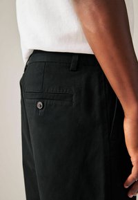 Zwarte broek van gladde stof, met een achterzak met knoopsluiting en een getailleerde taille. De broek is gecombineerd met een wit shirt.