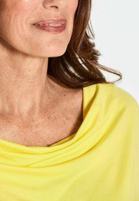 Gelb drapiertes Oberteil aus weichem Stoff, mit weitem Ausschnitt und glatter Textur, das auf nackten Schultern und dem Schlüsselbein sitzt.