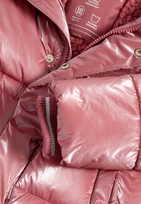Manga de chaqueta acolchada brillante de color rosa con puño de punto que muestra rayas grises y blancas, parcialmente desabrochada con forro de felpa rosa visible y símbolos de cuidado.