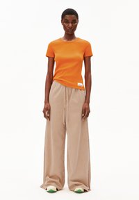 Oranje geribbelde top met korte mouwen gecombineerd met beige wijde broek en sneakers met groene accenten. Bevat een logo-opdruk op de top.