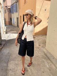 Femme portant un haut blanc sans manches, un short noir, des tongs rouges, des lunettes de soleil et un foulard sur la tête, marchant dans une rue étroite baignée de soleil.