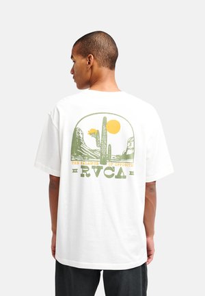 Uomo che indossa una t-shirt bianca con grafica di paesaggio desertico verde e giallo e sole con logo RVCA sulla schiena, in piedi davanti a uno sfondo semplice.