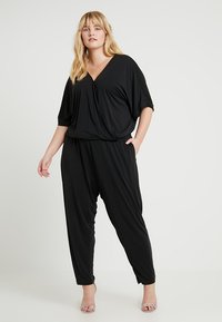 Svart jumpsuit med korte ermer og V-hals, med løs passform og sidelommer. Glatt stoff med drapert silhuett.