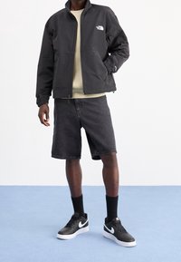 Chaqueta negra con cremallera y cuello alto, sudadera gris debajo, pantalones cortos de mezclilla negros, zapatillas negras y calcetines negros.