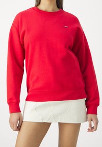 Femme portant un sweat-shirt Levi's manches longues rouge vif associé à une jupe courte blanche sur un fond uni.