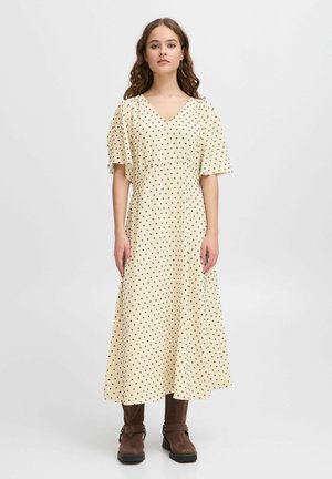 Beige mekko, jossa on pilkkukuvio ja V-mallinen kaula-aukko sekä lyhyet liehuvat hihat. Mallilla on päällään ruskeat korkokengät. Yksinkertainen tausta.