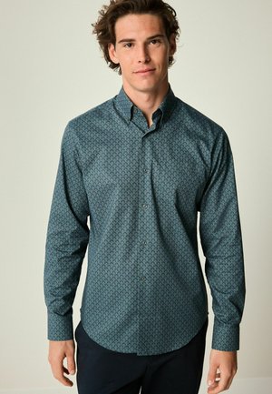 Blue-green button-up shirt with a scale pattern, long sleeves, and a rounded hem. Fabric appears smooth, with matching buttons.