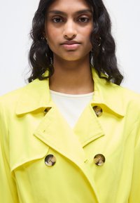 Κίτρινο παλτό τύπου trench με διπλό πέτο, μεγάλο γιακά και γυαλιστερές στρογγυλές κουμπότρυπες, φορεμένο πάνω από ανοιχτόχρωμο μπλουζάκι, με απαλή υφή.