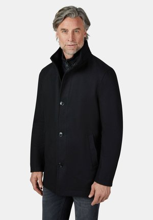 Mann mit grauem Haar und Bart, trägt einen schwarzen Knopfmantel und Jeans, steht vor einem schlichten hellen Hintergrund.