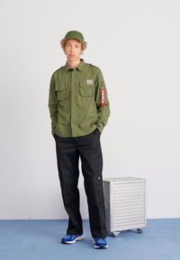 Green button-up shirt with two chest pockets, black trousers, and a matching cap. Silver rolling suitcase beside model. Blue sneakers on feet.