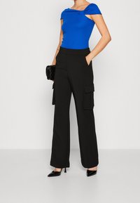 Top ajustado azul con diseño de hombros al descubierto, combinado con pantalones cargo negros de cintura alta con bolsillos laterales. Incluye tacones negros y un bolso.