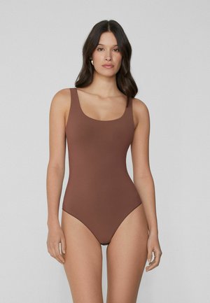 Femme aux longs cheveux noirs portant un maillot de bain une pièce marron sans manches, debout devant un fond gris clair uni.