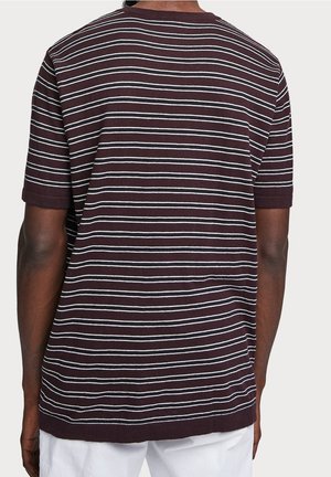 Uomo che indossa una T-shirt viola scuro a maniche corte con sottili righe bianche orizzontali, visto da dietro, abbinata a pantaloni bianchi.