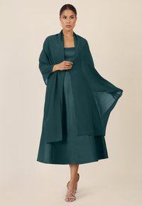 Türkises Satinkleid mit einem fließenden Überwurf, das ärmellos ist und einen knielangen Rock hat. Abgerundet wird das Outfit durch schlichte, silberfarbene Sandalen.