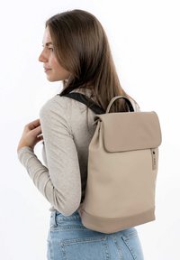 Zaino beige realizzato in materiale liscia, con chiusura arrotolata, spallacci neri regolabili e un sottile dettaglio di branding sul lato.