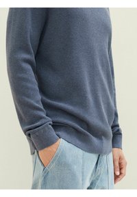 Pull en mélange de coton bleu foncé, avec un motif tricoté texturé et des poignets côtelés, associé à un jean en denim bleu clair.