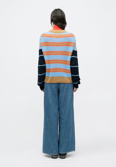 Πλεκτό πουλόβερ σε μπλε με πορτοκαλί, κόκκινες και καφέ ρίγες· μακριά μανίκια με σκούρες μπλε λεπτομέρειες· συνδυασμένο με τζιν παντελόνι φαρδιάς γραμμής.