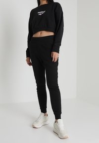 Sudadera negra corta con el logo de Reebok, combinada con joggers negros ajustados y zapatillas blancas con detalles en rosa claro.