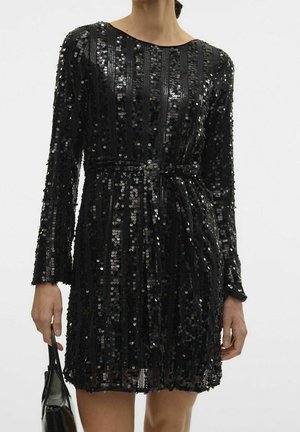 Woman wearing a black, long-sleeve sequin dress with a waist tie, holding a black handbag in her left hand against a plain background.